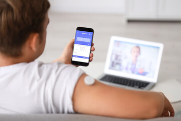 Diabetic young man with glucose sensor receiving online consultation from doctor at home, back view