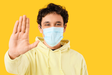 Young man in medical mask showing stop gesture on yellow background, closeup. Immunity concept