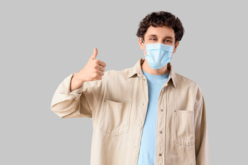 Young man in medical mask showing thumb-up on light background. Immunity concept