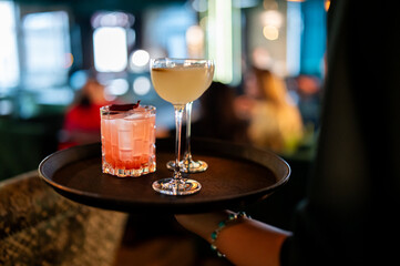 Two stylish cocktails, one pink and one yellow, are served on a dark tray in a dimly lit, upscale bar. Elegant evening drinks with a soft bokeh background