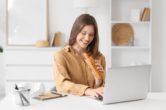 Young businesswoman with tasty hot dog working at desk in office