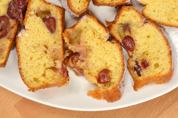 A close-up of several irregularly sliced pieces of baked yellow cake with visible dark red cherries and a dusting of powdered sugar on a white plate