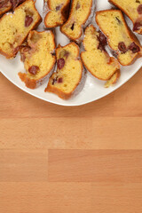 An overhead view of slices of baked yellow cake with cherries, dusted with powdered sugar, arranged on a white plate covering the top of a wooden background