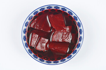An overhead view of deep red, pickled beetroot slices served in a glossy liquid within a small, blue and white patterned ceramic bowl on a white background