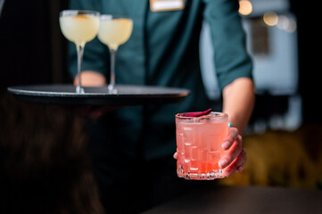 A vibrant pink cocktail with a garnish is hand-held in a dark, moody bar with other drinks on a tray.
