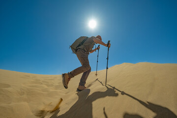 A hiker with a backpack and trekking poles ascends a sand dune under the bright sun. The clear blue sky above emphasizes the dynamic motion of the step in the desert.

