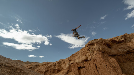 An adventurous traveler jumps across a canyon, frozen mid-air in a funny pose against a clear blue sky. Low-angle shot emphasizing motion, freedom, and playful challenge. © ANDREY