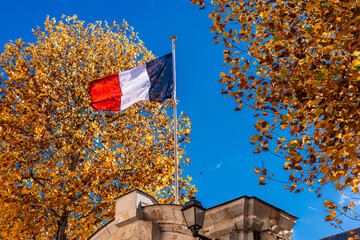 French flag waves proudly atop a stone structure, framed by vibrant autumn trees under a clear blue...