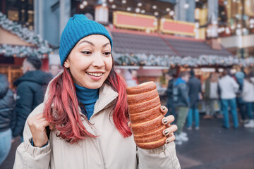 Young woman happily holding and looking at a chimney cake, enjoying traditional street food at a festive European Christmas market during winter