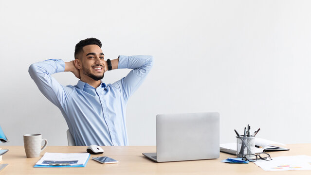 A young Arab man sits comfortably at his desk, smiling and leaning back in his chair. He feels satisfied after completing his tasks for the day, enjoying a moment of relaxation.