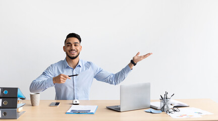 Young Middle Eastern businessman sits at a desk, smiling and pointing to a free space as if inviting ideas or advertisements. His cheerful demeanor creates a positive work atmosphere.