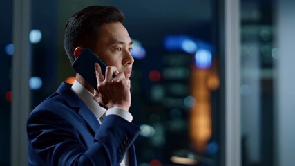 A businessman in a formal suit stands in a dimly lit office while on the phone, city lights glowing in the background - Powered by Adobe