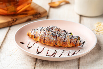 Freshly baked croissant topped with chocolate drizzle and powdered sugar served on a pink plate