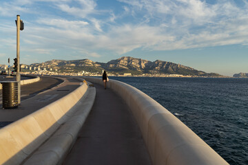 A woman walks on the Corniche, the famous seaside promenade in Marseille, France, overlooking the Mediterranean Sea and city mountains.