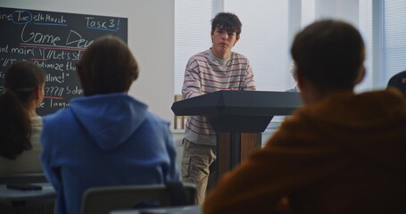 Teenage Boy at Podium Explains Work Peers, Game Dev Topic Visible on Blackboard. Concept Computer Science Education, Process of Peer Review and Discussion in Modern, Technology Focused Classroom.