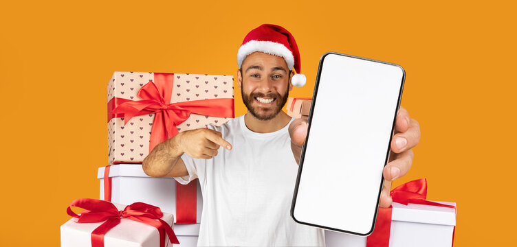 A man wearing a Santa hat smiles brightly while holding a smartphone in front of colorful gift boxes stacked high. The scene radiates holiday cheer and excitement for the season.