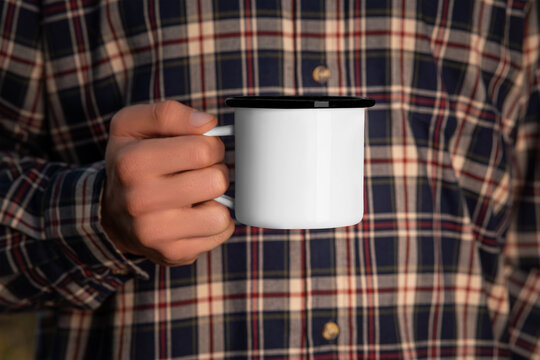 Mockup of a white metal mug with a black rim in the hand of a man in a flannel shirt.