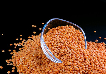 Red lentils in a transparent glass cup on a black background. Side view. Lots of lentils around the cup.