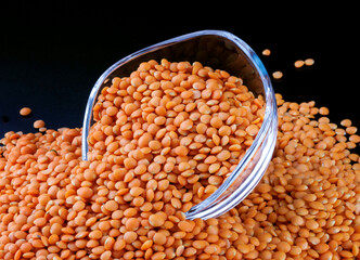 Red lentils spilling out of a glass cup on a black background. Side view. Lots of lentils around the cup.