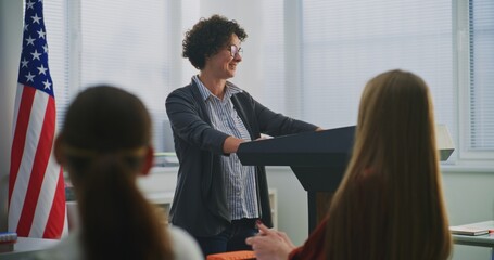 American Teacher Stands Podium Beside US Flag, Engaging Students In Open Discussion About National...