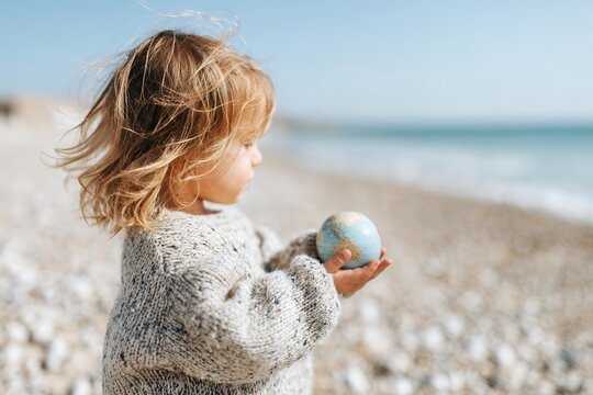 Child holding globe on sandy beach looking at the ocean waves