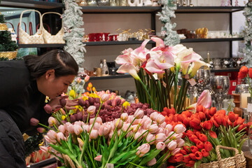 A woman leans over to smell a variety of colorful flowers arranged on a table in a florist shop. The background features shelves with decorative items and holiday ornaments.