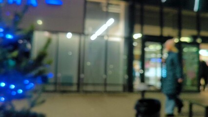 A woman walks along an evening street decorated for Christmas. Blurred. Along the storefronts of trendy shops and a cinema along evening street in blurred focus. Before Christmas