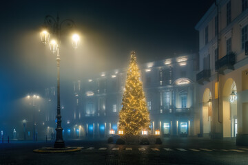 A brightly lit Christmas tree stands in a foggy, empty city square in Asti, Italy.