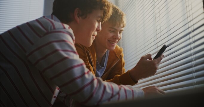 Teen Boys by Bright Classroom Window Scroll Through Phone Together, Exchanging Quiet Comments. Concept of Technology in Youth Culture, Casual Communication, Positive Emotions and Friendship Moments.