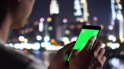 Close up of a person holding a smartphone with a green screen interacting with the device against a blurred backdrop of a vibrant city skyline at night showcasing modern technology and urban connecti. - Powered by Adobe
