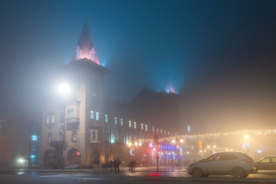 A foggy evening scene of a beautiful historic building illuminated with Christmas lights, with people walking and a car parked in the foreground. Saratov Conservatory in Mist foggy night lighting.