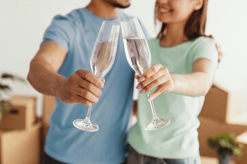A young couple stands together in a cozy room surrounded by moving boxes, joyfully toasting with glasses of sparkling water. They smile at each other, celebrating a new beginning.