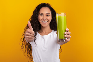 Brightly lit studio features a young woman with curly hair happily holding a green smoothie. She shows a thumbs up, promoting a healthy lifestyle and enjoying her nutritious drink.