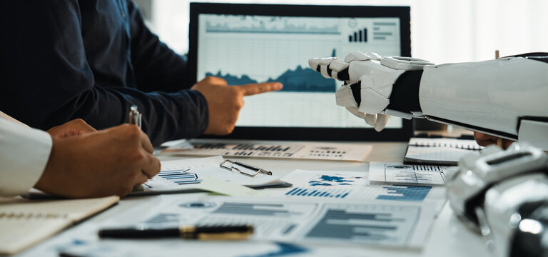 Human and robotic hands work together in a modern office, analyzing data on a computer screen filled with charts and graphs, symbolizing innovative collaboration.Noogenesis - Powered by Adobe