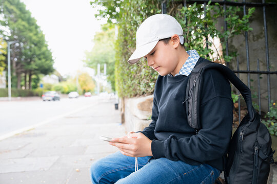 Teenage boy using smartphone sitting on city sidewalk,wearing casual clothes and backpack waiting for bus travel to school,Concepts digital lifestyle,Mobile communication,Student life,Urban tech usage