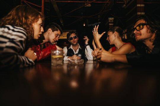 A lively group of friends sits around a bar, chatting, laughing, and enjoying drinks late at night. One man wears sunglasses and a vest, adding a stylish vibe to the gathering.