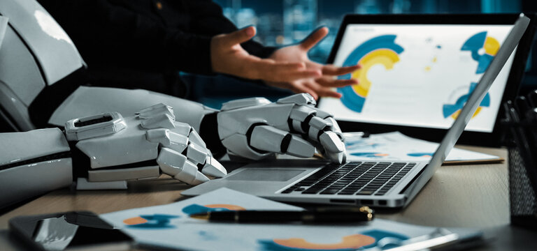 A human-like robotic arm gestures dynamically over a laptop in a modern office setting, highlighting the blend of technology and business in today's workspace.Noogenesis