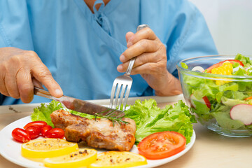 Asian senior woman patient eating pork chop stake and vegetable salad for healthy food in hospital.