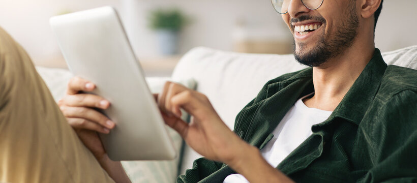 A man relaxes on a sofa, smiling as he interacts with a tablet. His casual attire and the comfortable setting suggest a moment of joy and convenience.