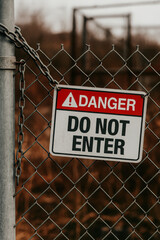 Danger “Do Not Enter” warning sign attached to metal fence with a chain, set outdoors against a blurred industrial background. A clear message of safety, restricted area, and hazard prevention