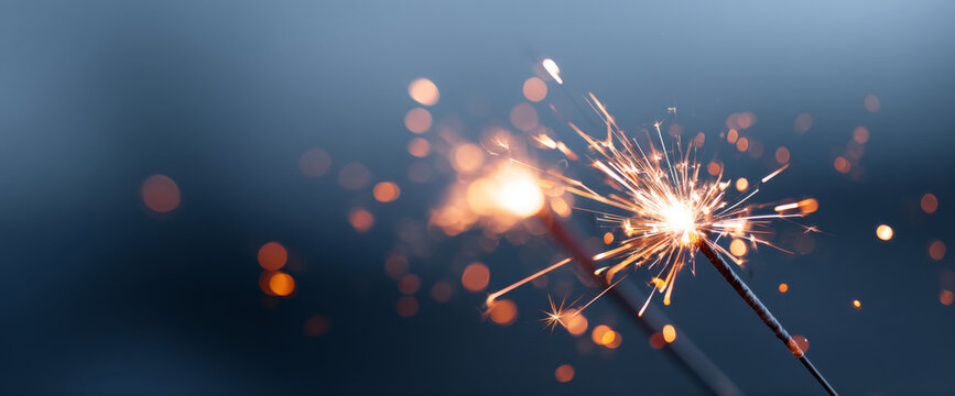 Close-up of lit sparklers emitting bright sparks with bokeh light effects against a dark blue background - Powered by Adobe