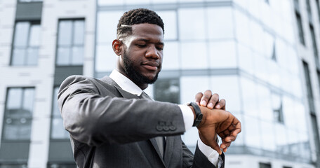 A man dressed in a formal suit is checking his watch while standing in front of a contemporary office building. The scene captures a moment of waiting or planning in an urban environment.