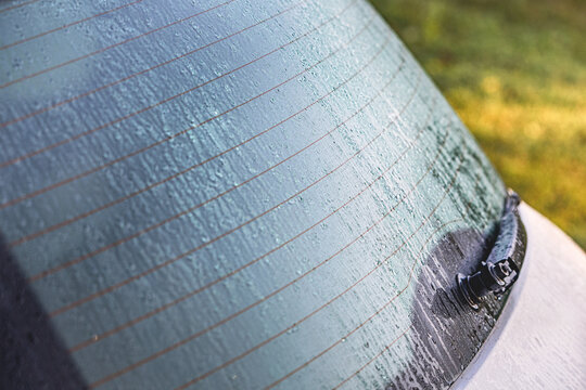 Car rear window covered with rain drops and defroster lines. Wiper resting on wet glass
