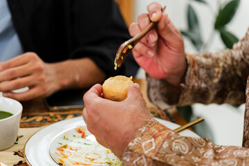Close-up of diners enjoying Indian street food, with a hand filling a pani puri using a spoon beside a plate and a bowl of green chutney at a table.