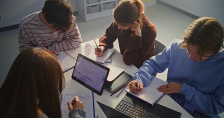 Diverse Study Group of Young Men and Women Leaning Over Table, Engaged in Academic Work. Student Various Study Methods Employed, Digital Screen, Handwritten Notes, Modern Study Environment. Top View.