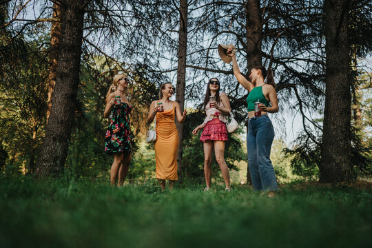 A lively outdoor scene in a green park shows five young women socializing, laughing, and toasting drinks. They wear colorful summer outfits, celebrating friendship and a carefree afternoon in nature. - Powered by Adobe
