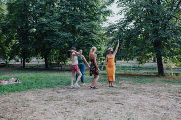 Obraz premium A group of friends enjoy a playful dance in a grassy park under leafy trees. One woman in a bright orange dress leads, while others smile, chat, and soak up warm summer vibes.