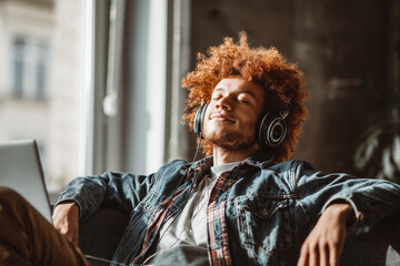 Young man wearing headphones relaxing on a couch in warm sunlight — cozy lifestyle portrait of music, leisure, and contentment.