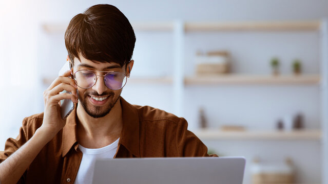 A young man sits at a desk, smiling while engaged in a phone call. He is using a laptop, surrounded by a bright and tidy workspace decorated with minimalistic items.