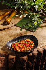 A vibrant mix of tomatoes, herbs, and spices arranged beautifully in a black dish sits on a rustic wooden table, accented by olive branches and kitchen tools in the background.